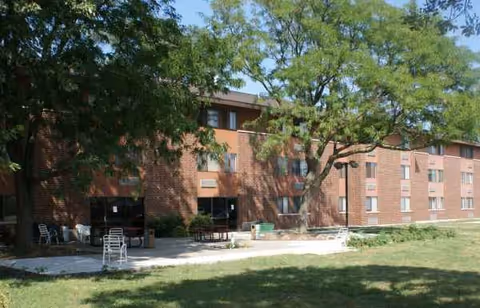 Brick senior apartment building with trees, lawn, and outdoor seating.