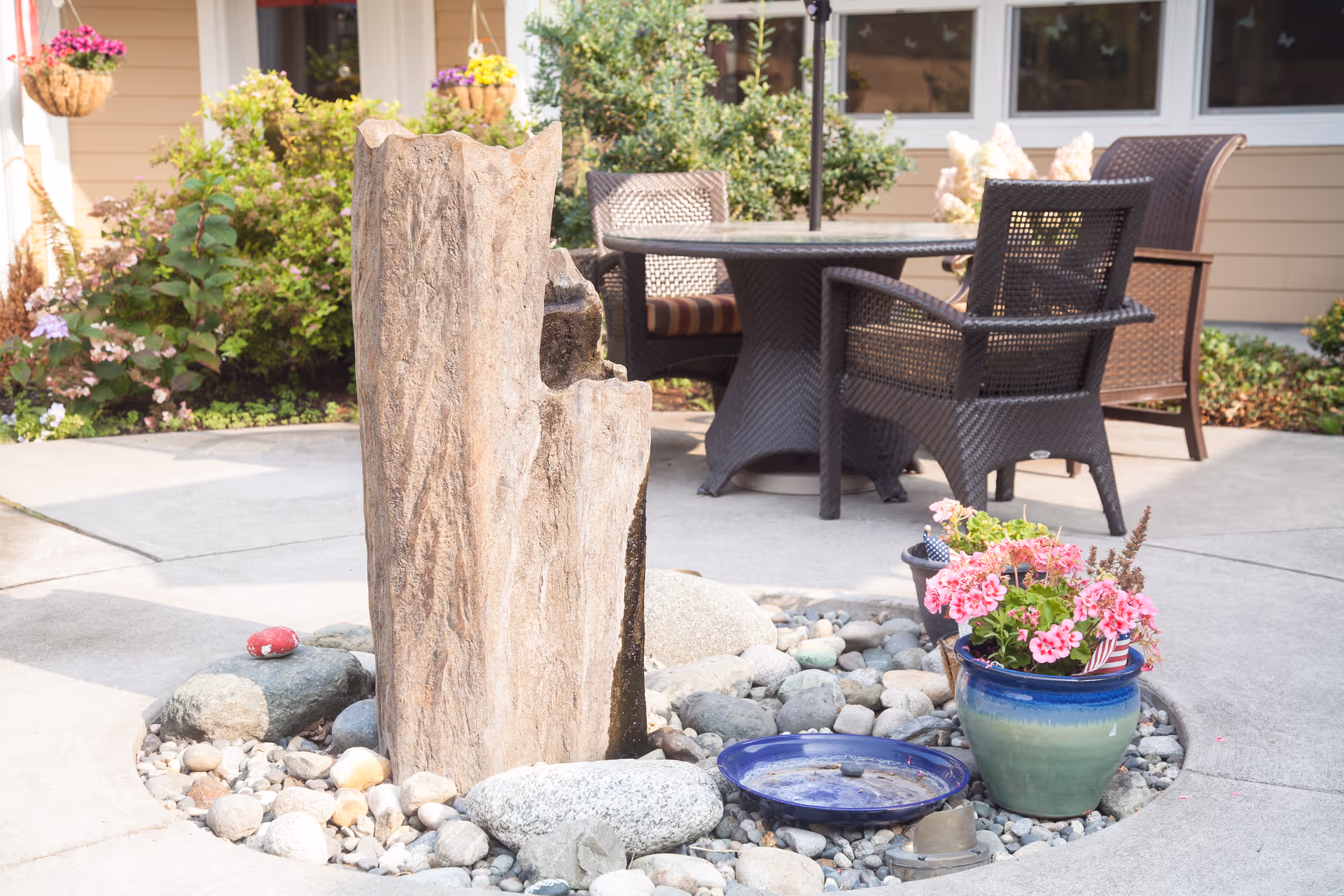 Sunlit courtyard patio with a central rock-and-wood fountain, potted flowers, and wicker dining chairs near a building.