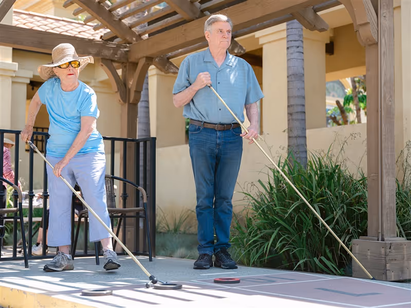 An elderly woman and man playing shuffleboard outdoors under a wooden pergola. The woman is wearing a wide-brimmed hat, sunglasses, a light blue shirt, and light-colored pants, while the man is dressed in a blue short-sleeve shirt and jeans. They are focused on the game with shuffleboard discs on the court.