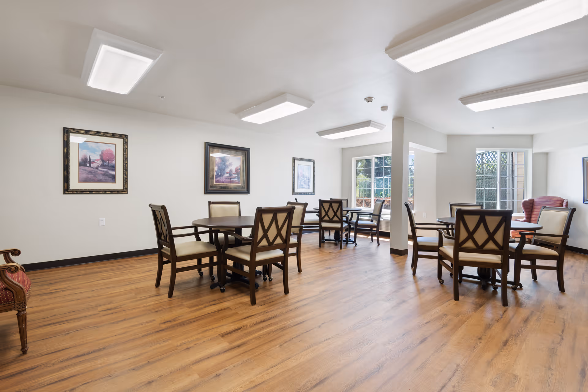 Bright communal dining/activity room with multiple round tables and chairs, wood floors, framed artwork, and large windows.