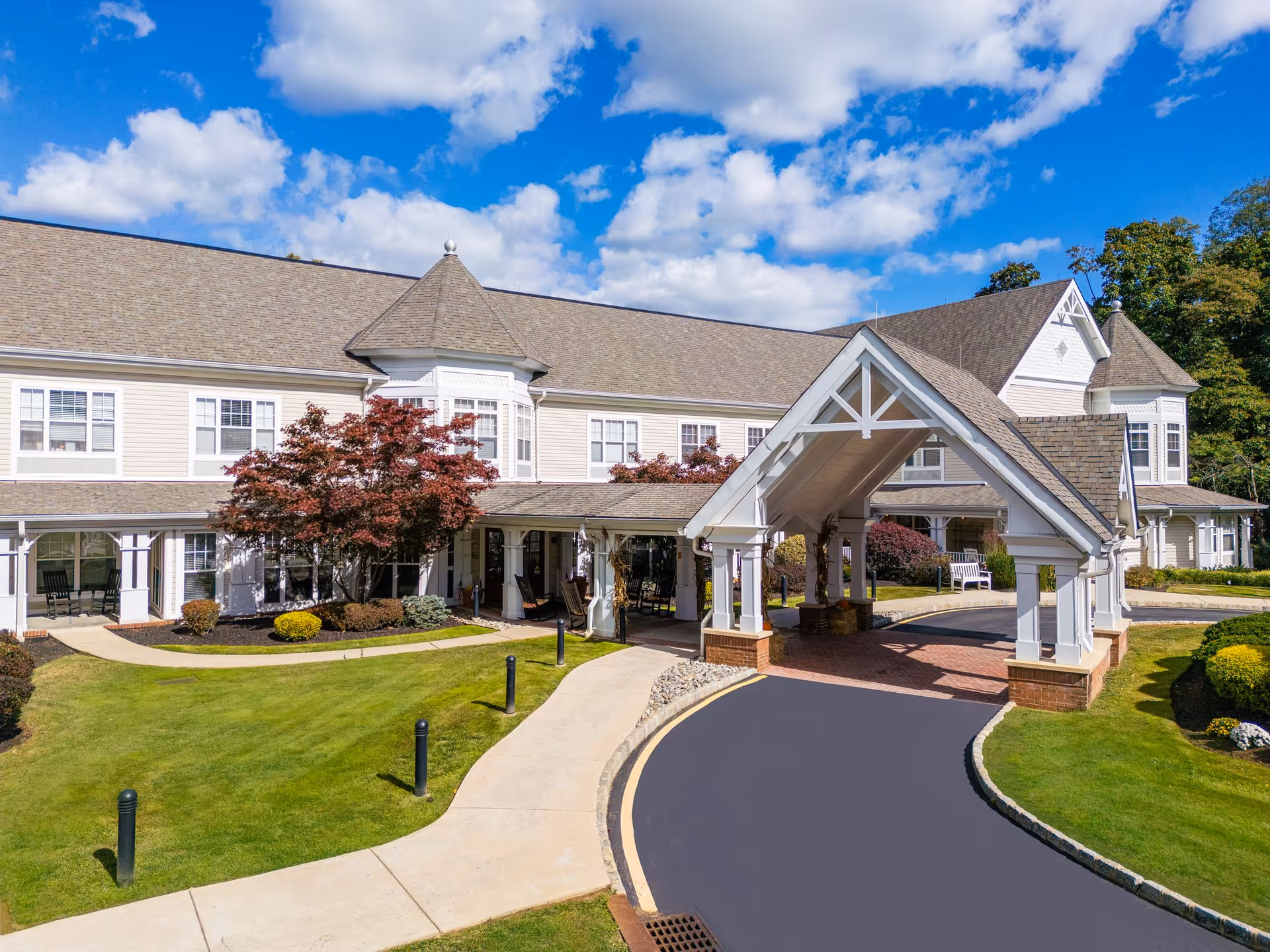 Exterior view of a senior living facility building with a covered entrance driveway, well-maintained green lawn, shrubs, and trees under a partly cloudy blue sky.