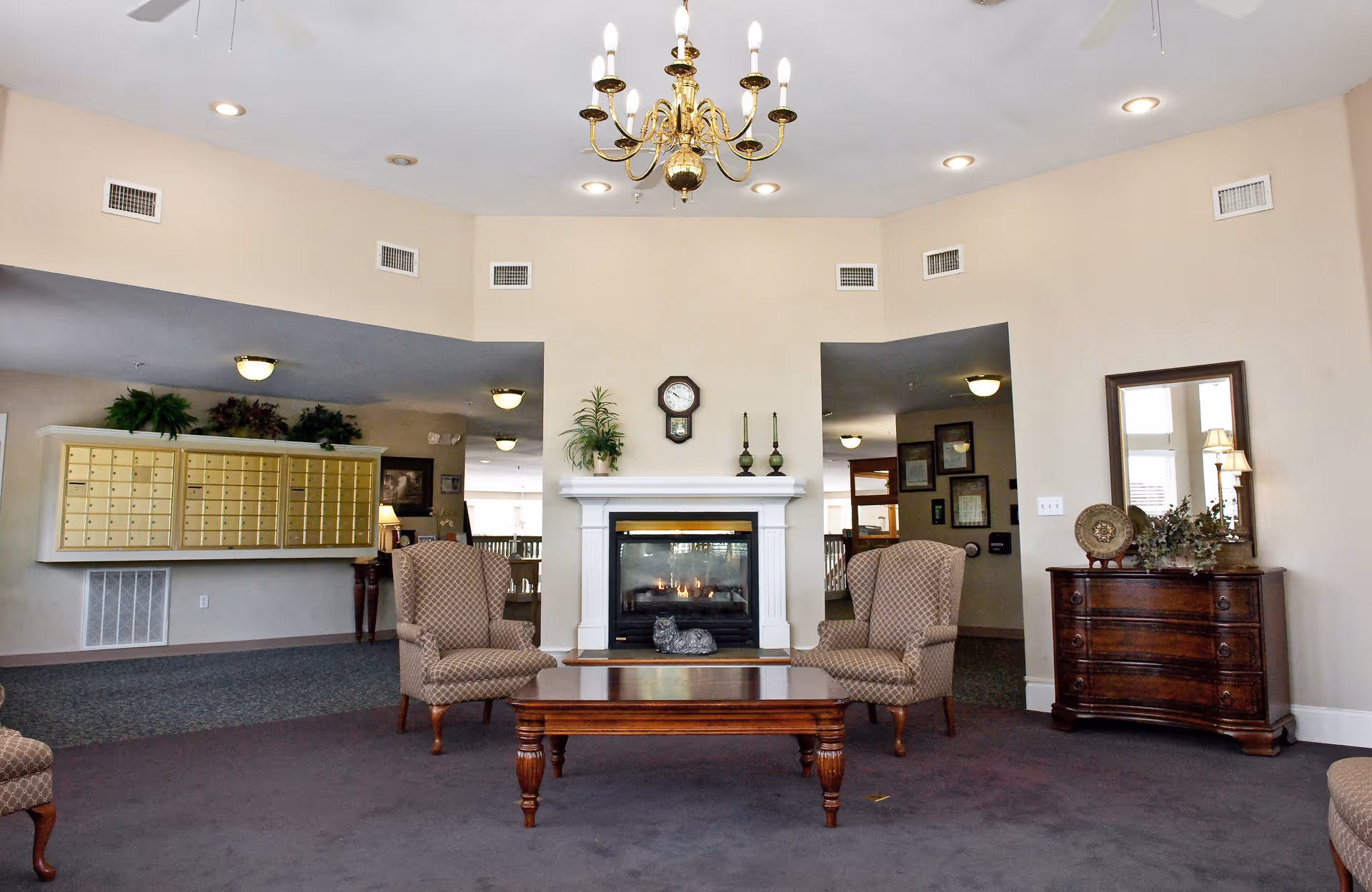 A cozy assisted living facility common area with a fireplace in the center, flanked by two patterned armchairs and a wooden coffee table. Above the fireplace is a wall clock and decorative items. To the right is a wooden dresser with a mirror and plants, and to the left are mailboxes mounted on the wall. The room has beige walls, carpeted floor, ceiling fans, and a chandelier.