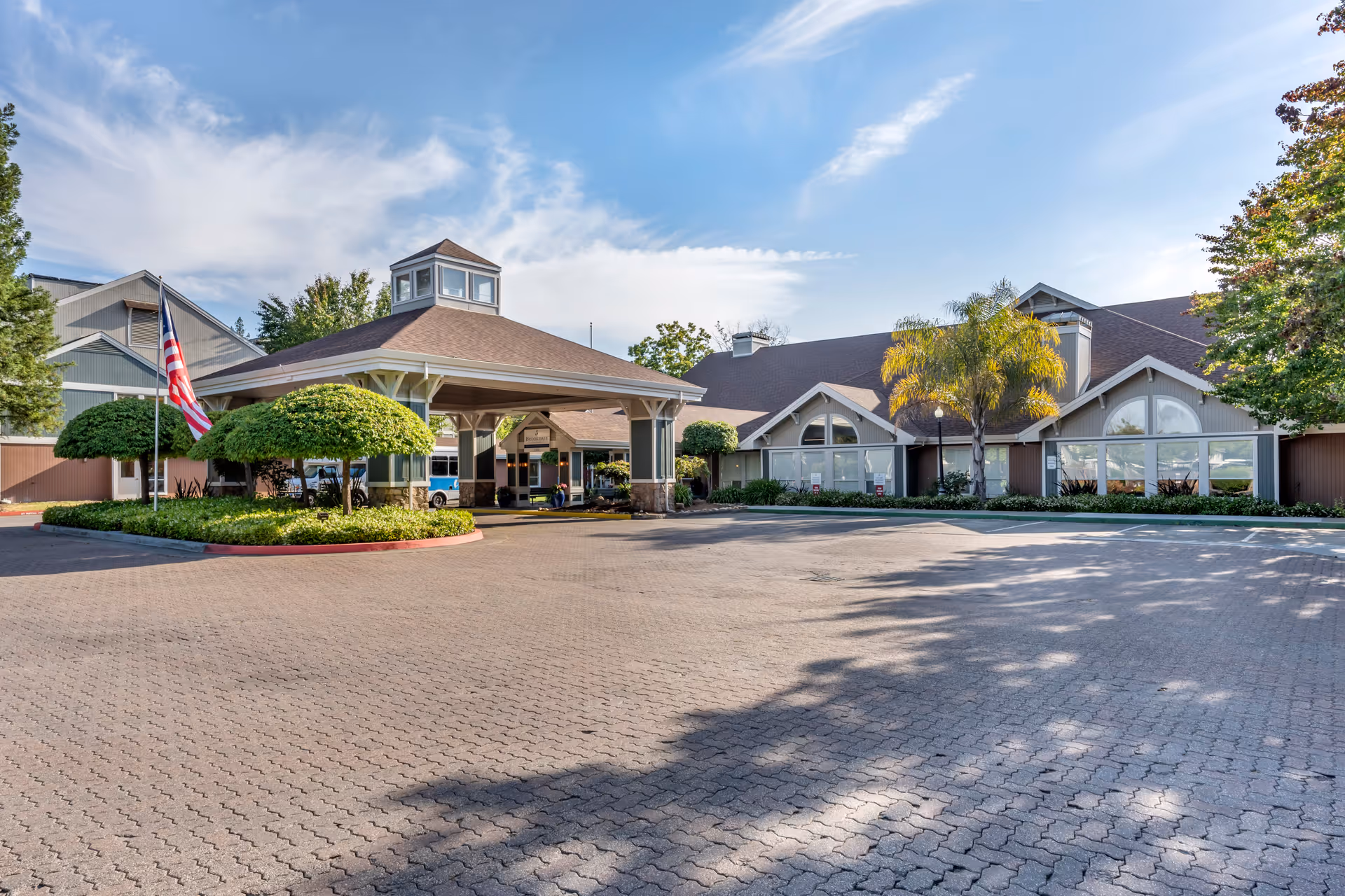 Exterior view of Brookdale Paulin Creek senior living facility showing a large paved driveway, a covered entrance with a cupola, landscaped greenery including trimmed bushes and a palm tree, and a clear blue sky with some clouds.