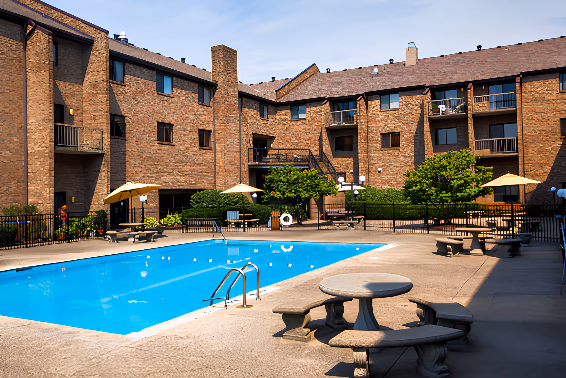 Outdoor swimming pool area at Brookwood Retirement Community with blue water, surrounded by concrete deck, stone tables with curved benches, yellow umbrellas, and a multi-story brick building in the background.