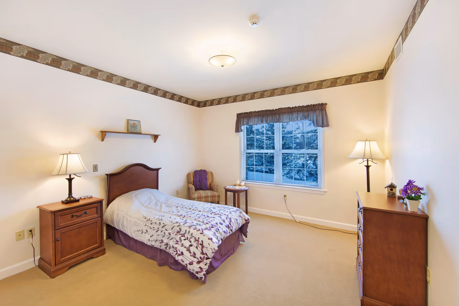 A cozy bedroom with a single bed featuring a white and purple floral bedspread. There is a wooden nightstand with a lamp on the left side of the bed, a small wooden dresser with a lamp and a purple flower pot on the right side of the room, and a striped armchair with a purple throw blanket near a window with blinds and a purple valance. The walls are light-colored with a decorative border near the ceiling.