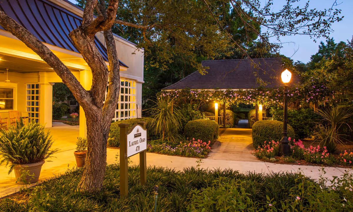Evening view of an outdoor garden area at Fellowship Brookside Assisted Living Community featuring a lit pathway under a wooden pergola covered with flowering vines, surrounded by bushes and flowers. A tree and potted plants are near a building entrance with warm lighting and rocking chairs.