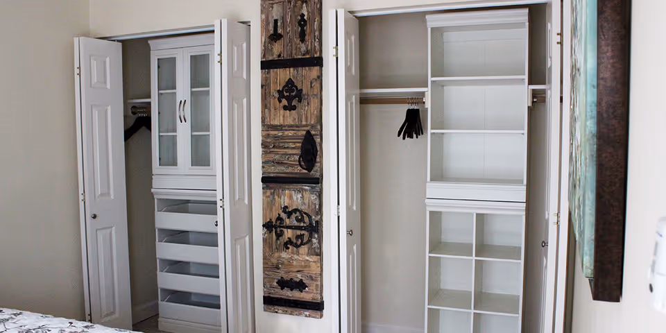 Interior view of a bedroom closet area with white bi-fold doors open to reveal shelving units and hanging rods with hangers. A decorative wooden panel with black metal accents is mounted on the wall between the closets. Part of a bed with a patterned bedspread is visible in the foreground.