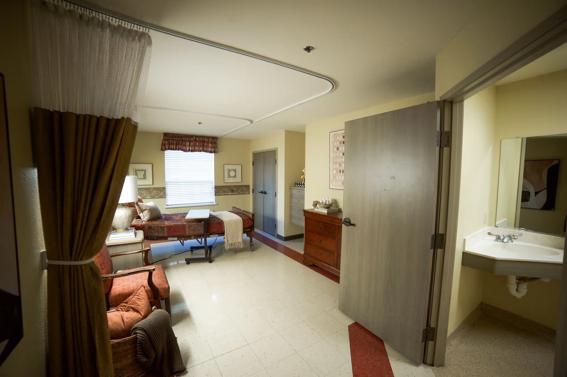 A tidy assisted-living bedroom with a bed, armchair, dresser, and an open door showing a sink area.