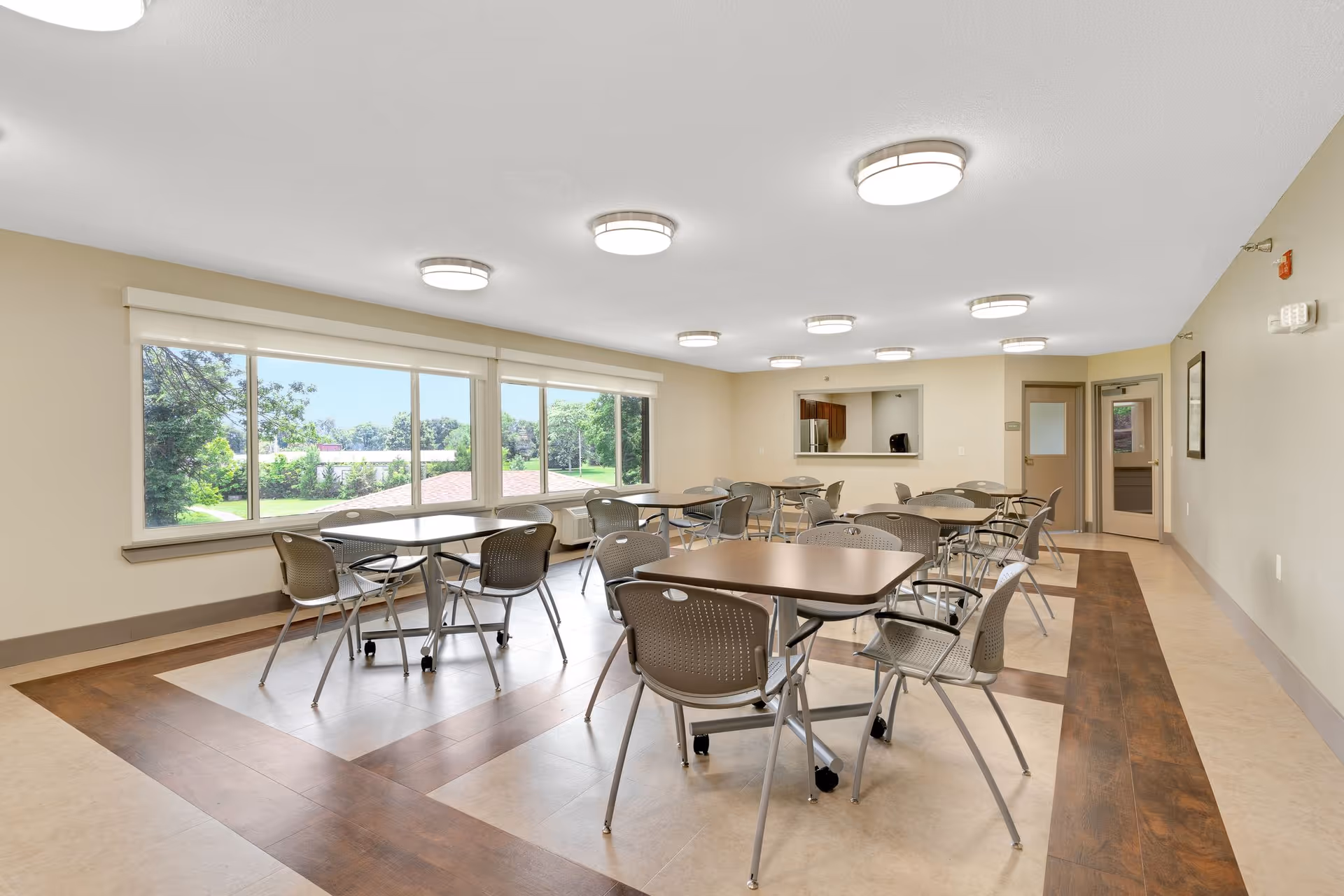 A bright dining room with multiple tables and chairs arranged neatly. Large windows on one side provide a view of green trees and a sunny outdoor area. The room has beige walls, a patterned floor with wood accents, and ceiling lights evenly spaced.