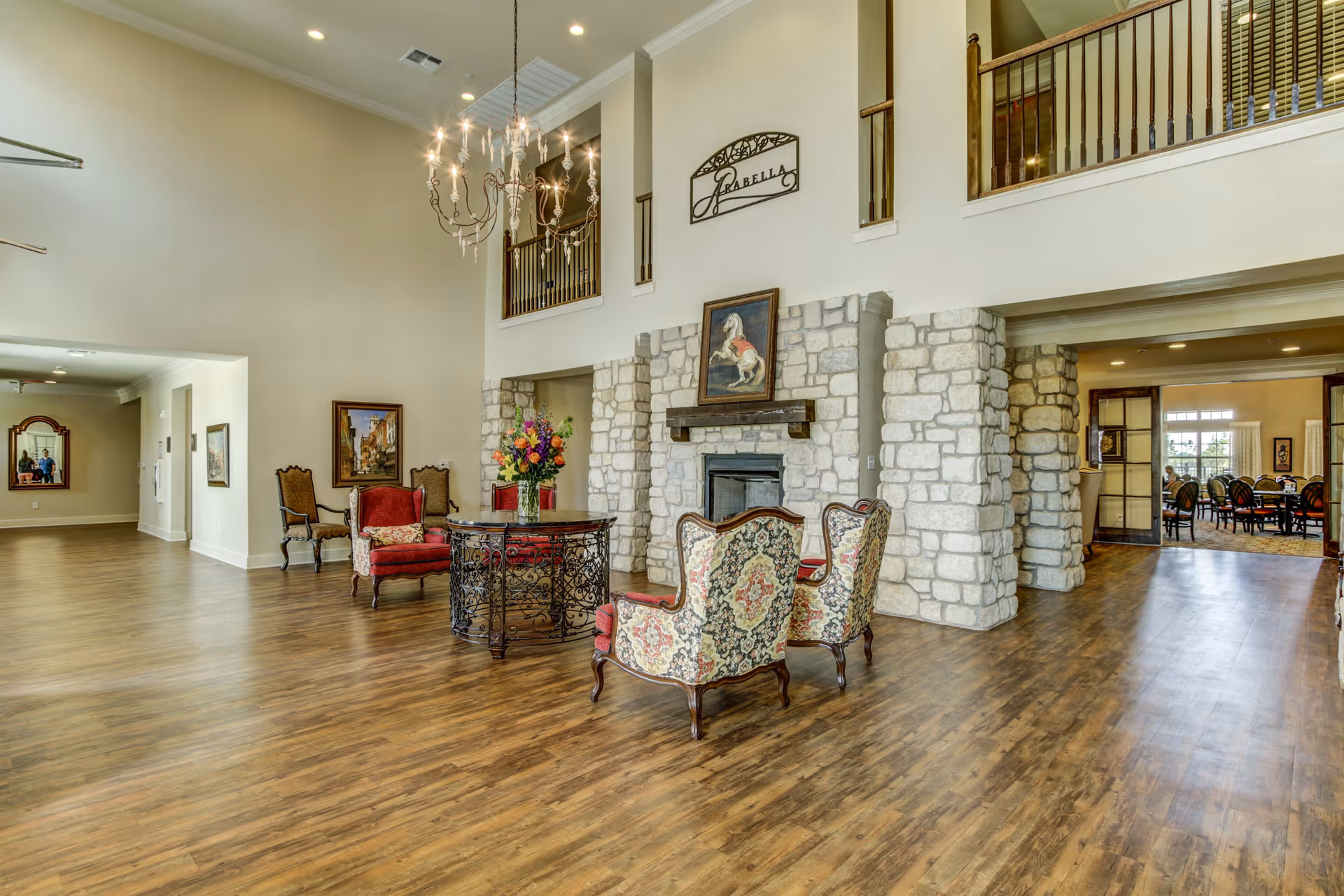 Spacious senior living facility common area with high ceilings, a chandelier, stone fireplace, and elegant vintage-style chairs arranged around a round table with a flower vase. The room features wood flooring, framed artwork on the walls, and a balcony railing above. In the background, a dining area with tables and chairs is visible through an open doorway.