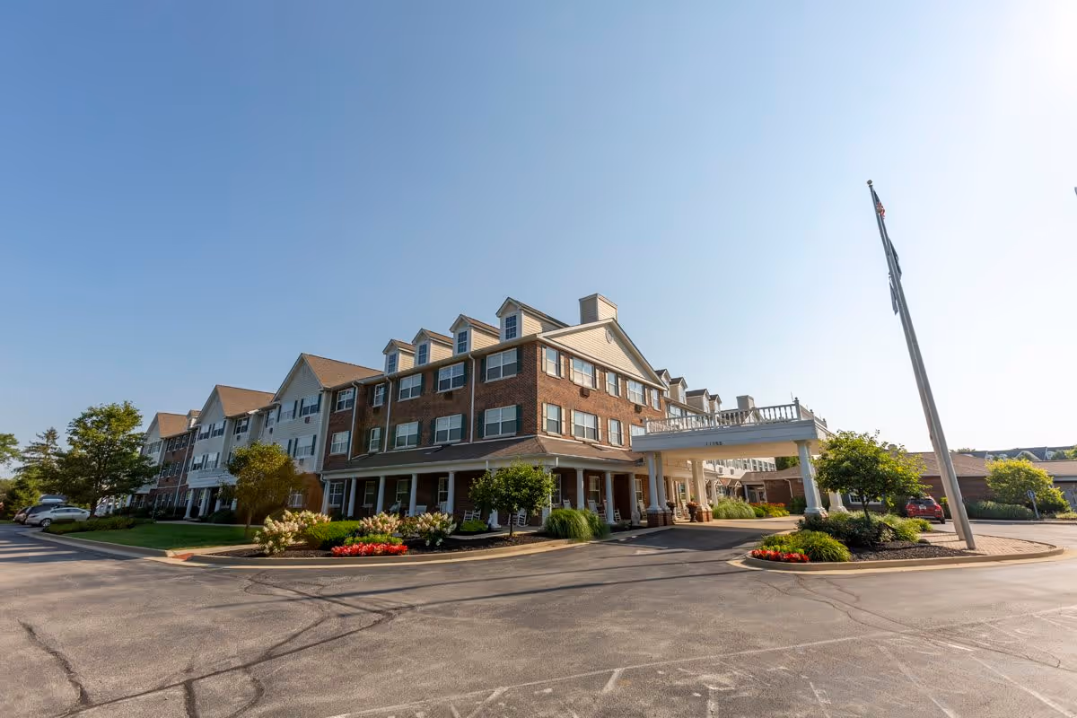 Exterior view of a large, multi-story senior living facility building with a covered entrance, landscaped flower beds, and a flagpole with an American flag. The sky is clear and blue.