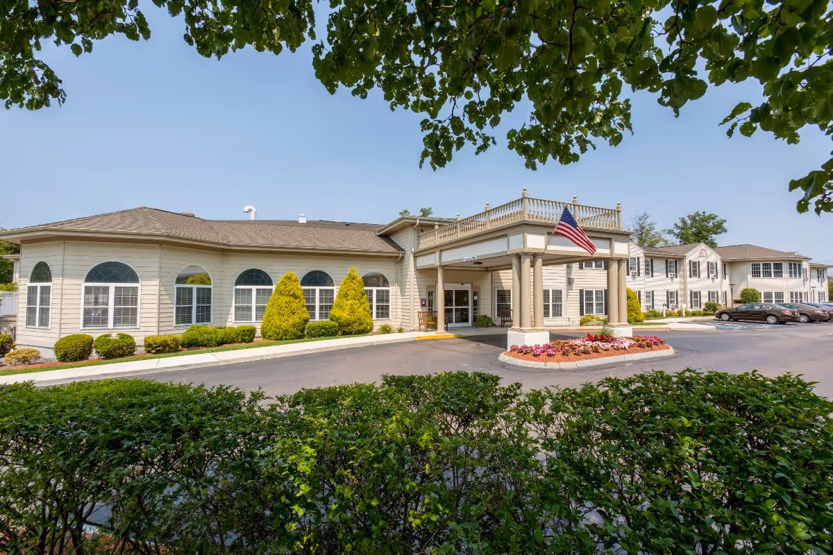 Exterior view of Benchmark Senior Living at Waltham Crossings showing a beige building with multiple windows, a covered entrance with columns, an American flag, landscaped bushes and flowers, and a parking area with cars under a clear blue sky.