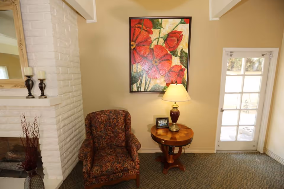 A cozy corner in a living room featuring a patterned armchair next to a round wooden side table with a lamp and a small framed picture. Above the table hangs a colorful painting of red flowers. To the left is a white brick fireplace with two candle holders on the mantel, and to the right is a glass-paned door leading outside.