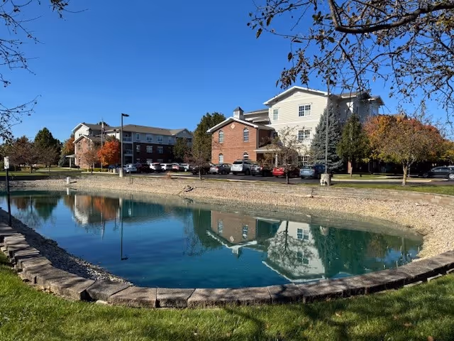 A senior living facility building named The Waterford at Levis Commons is visible behind a small pond with clear reflections of the building and trees. The scene is set on a sunny day with a clear blue sky and some autumn-colored trees around the parking area.