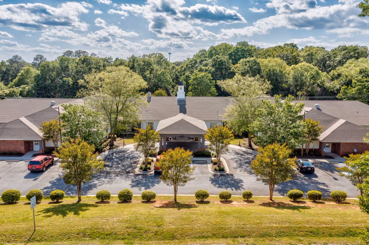 Aerial view of TerraBella Marchbanks facility showing a single-story building with a covered entrance, surrounded by trees and greenery under a partly cloudy sky. There are parked cars and a driveway in front of the building.