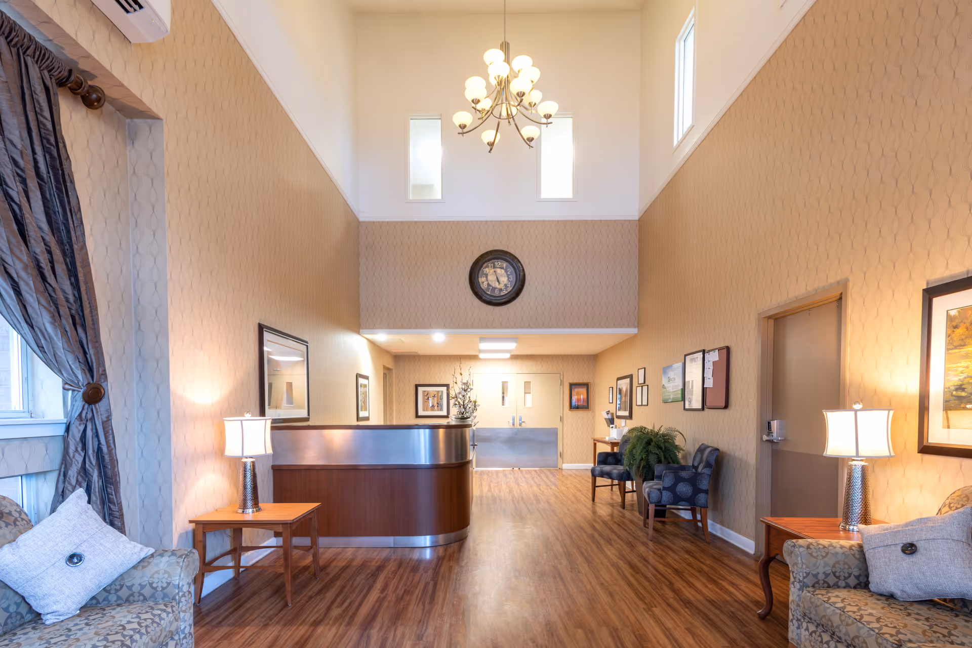 A spacious and well-lit reception area with high ceilings, featuring a wooden reception desk, two armchairs with patterned upholstery, side tables with lamps, framed artwork on the walls, a large clock above the reception desk, and windows with curtains allowing natural light.