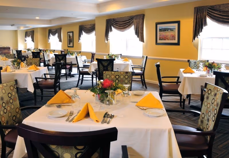 Bright dining room with neatly set tables, yellow napkins, patterned chairs, and floral centerpieces.