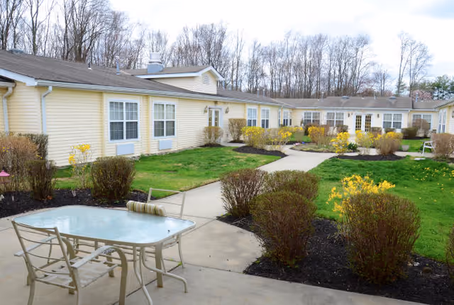 Outdoor patio area at Granville Place with a glass-top table and metal chairs on a concrete surface, surrounded by landscaped bushes and yellow flowers. A single-story yellow building with multiple windows and doors is visible in the background, along with leafless trees.