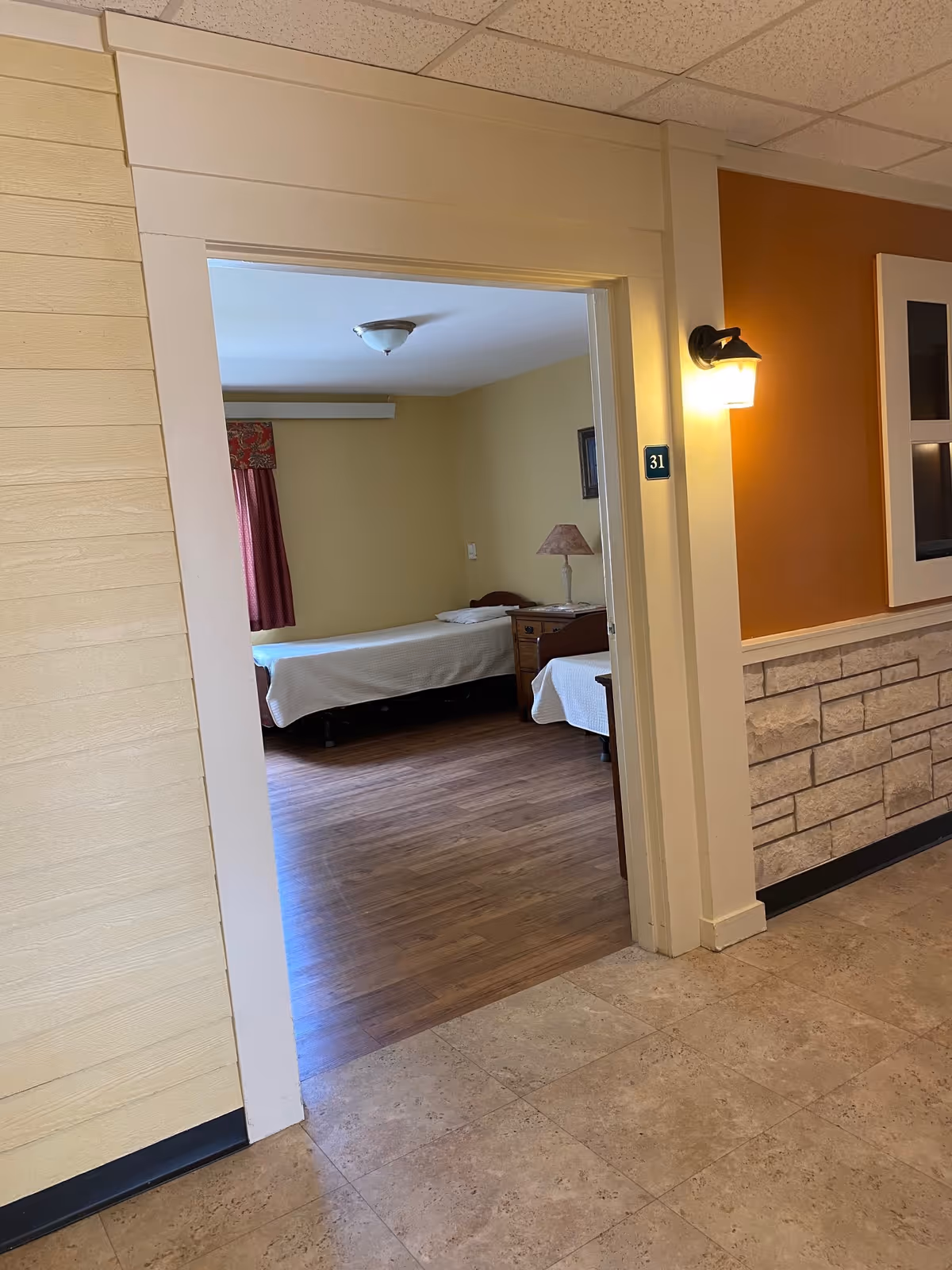 Open doorway from a corridor into a resident bedroom with two single beds, a nightstand and lamp and room number 31 on the frame.