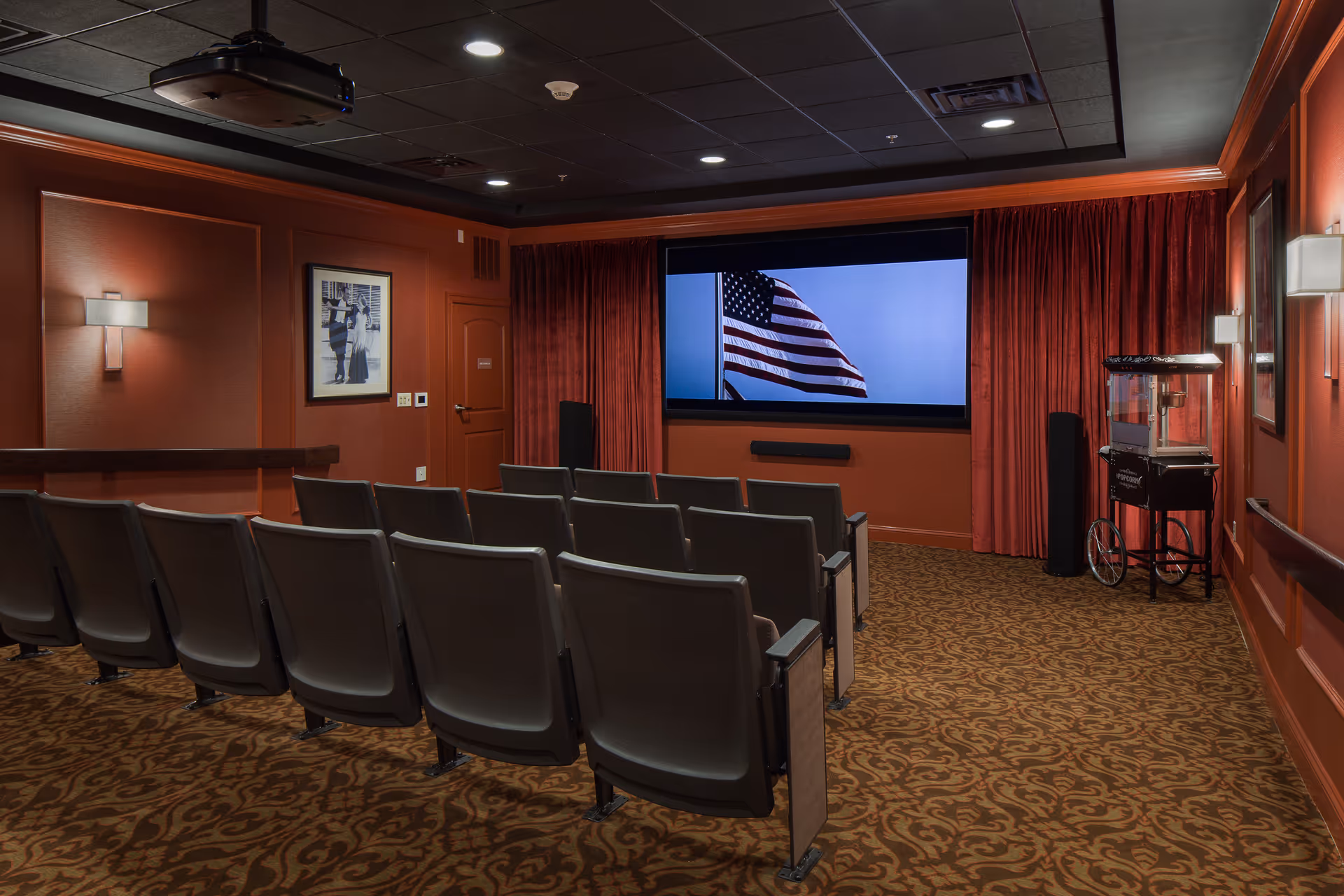 A small theater room with rows of gray seats facing a large screen displaying an American flag. The room has red walls, patterned carpet, wall sconces, a popcorn machine on the right side, and a ceiling-mounted projector.