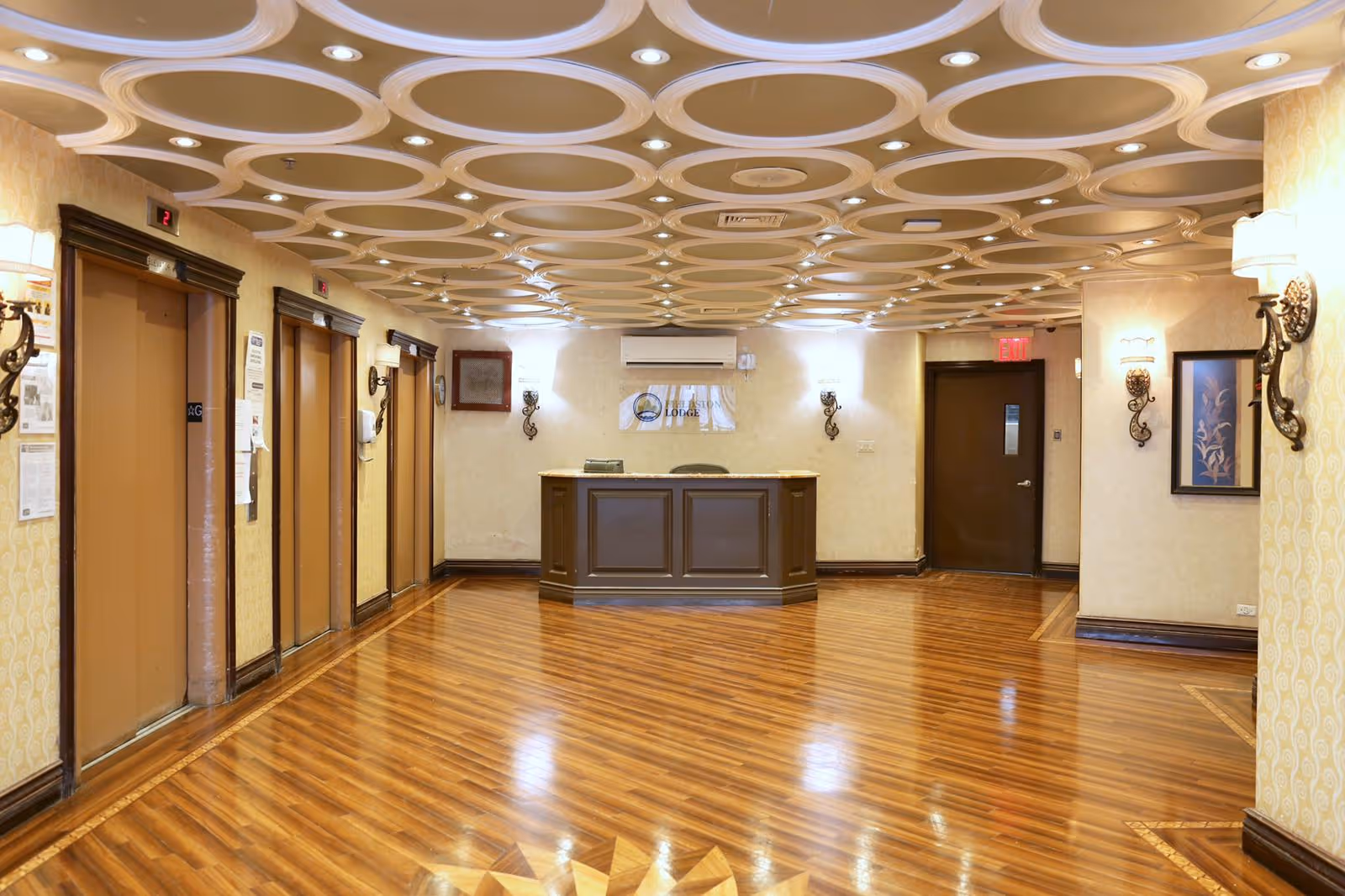 Interior view of a facility lobby area with polished wooden floors, a decorative ceiling with circular patterns, three elevators on the left side, a reception desk in the center back wall, wall sconces with lights, framed artwork, and an exit door on the right.