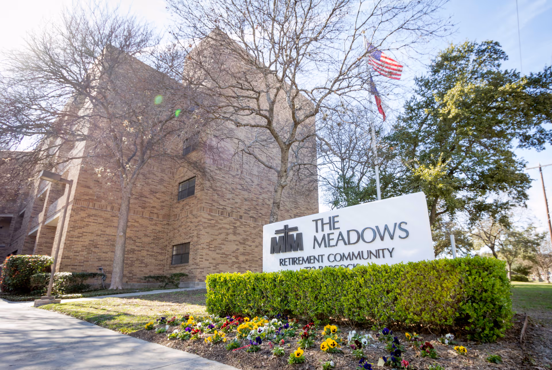 Exterior view of a brick retirement community building with a sign reading 'The Meadows Retirement Community', a flowerbed, and an American flag.