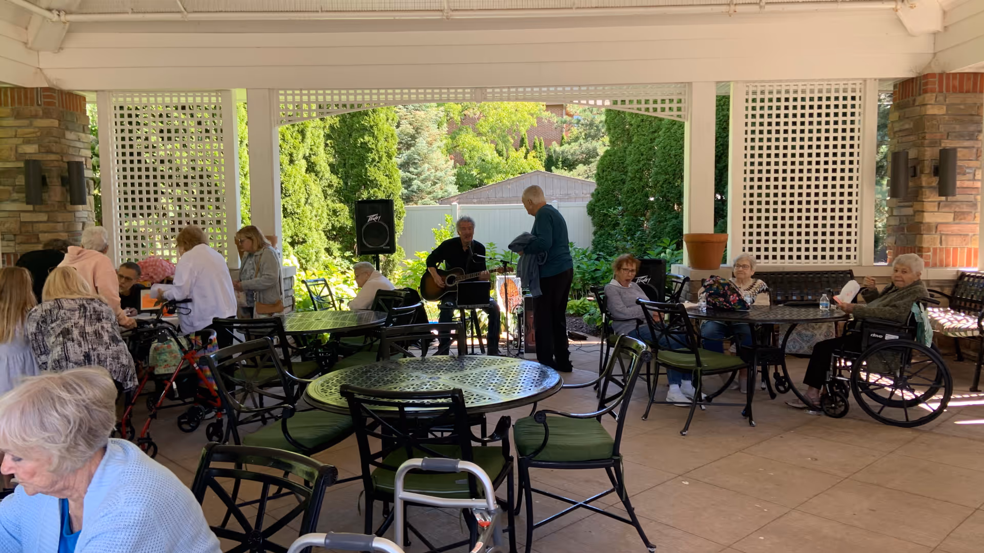 Seniors gathered and seated at tables on a covered outdoor patio while a musician plays guitar.