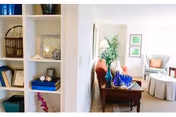 Bright and airy interior space featuring a white shelving unit with books and decorative items on the left, a narrow wooden table with a blue glass vase and a lamp in the center, and a cozy seating area with a white armchair, a round ottoman, and framed artwork on the wall in the background.