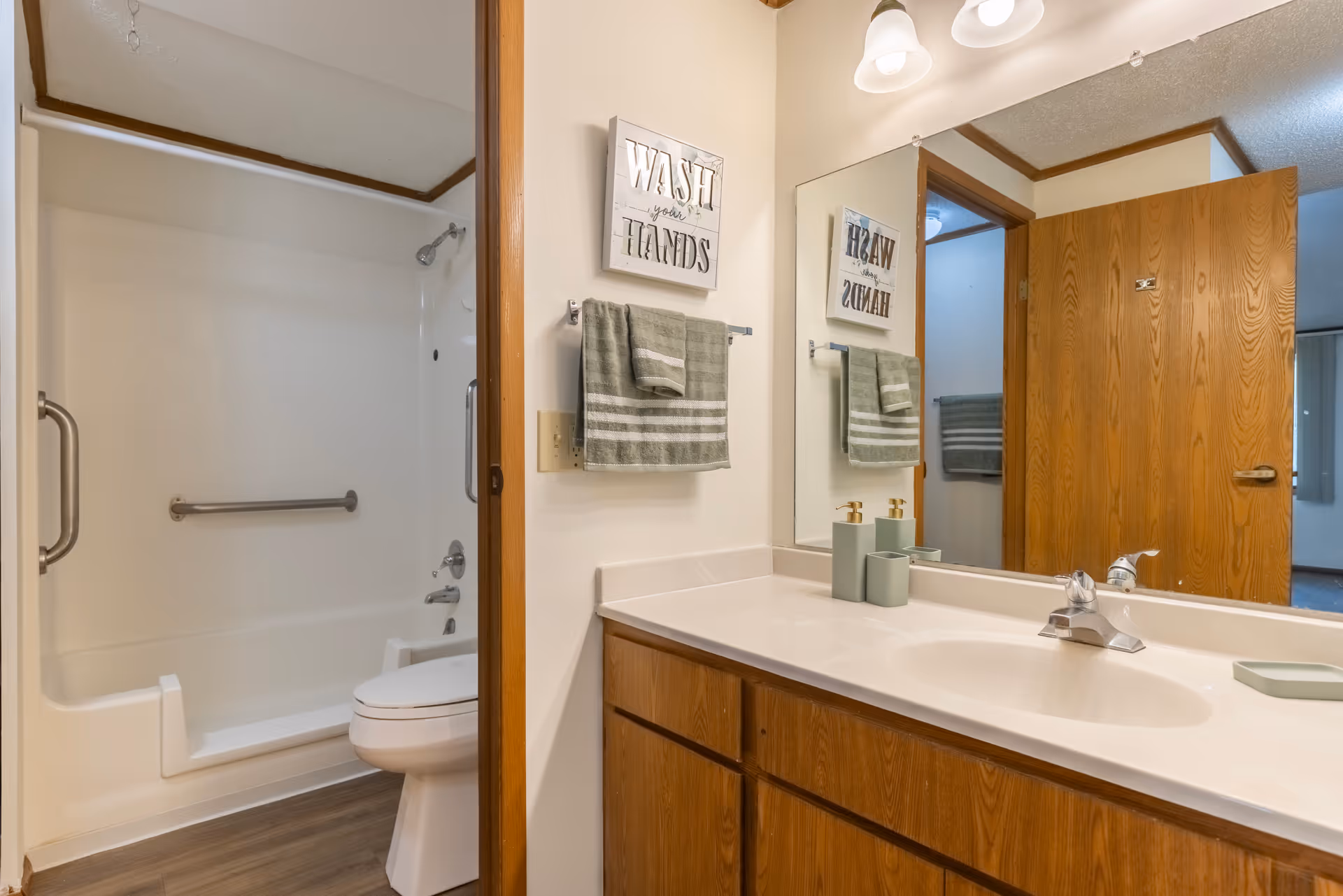 A bathroom with a white sink and countertop, wooden cabinets underneath, a large mirror above the sink, and a wooden door. To the left, there is a shower and bathtub combination with grab bars and a toilet. A towel rack with a green towel and a sign that reads 'WASH your HANDS' are mounted on the wall.