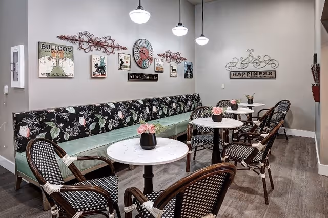 A cozy seating area with a long floral-patterned bench along the wall and several round white tables with black bases. Each table has a small black vase with pink flowers. There are black and white woven chairs around the tables. The wall is decorated with various framed pictures, a large clock, and decorative metal wall art. The floor is wood, and three pendant lights hang from the ceiling.