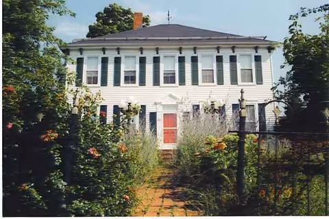 Two-story white house with green shutters, red door, and a garden path.