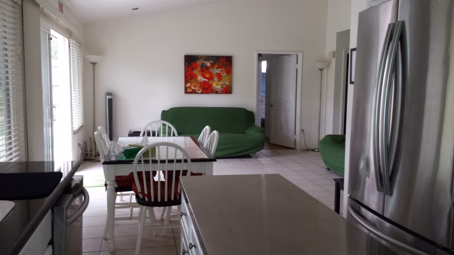 Interior view of a living and dining area with a kitchen island in the foreground. There is a stainless steel refrigerator on the right, a dining table with white chairs and red cushions in the middle, and two green sofas against the far wall. A colorful floral painting hangs on the wall above one sofa. Large windows with blinds are on the left side, letting in natural light.