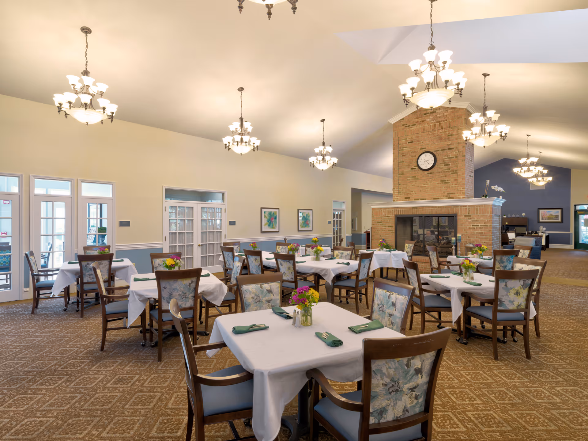 A spacious dining room with multiple tables covered in white tablecloths, each set with green napkins and small flower arrangements. The room features a large brick fireplace with a clock mounted above it, elegant chandeliers hanging from the ceiling, and floral upholstered chairs. The walls are painted in light colors with some framed artwork, and there are glass doors leading to another area.