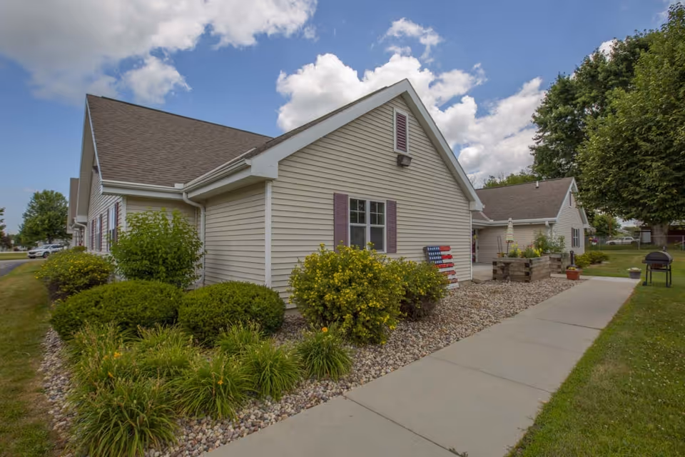 Single-story beige assisted living building with shrubs, a walkway, and a partly cloudy sky.