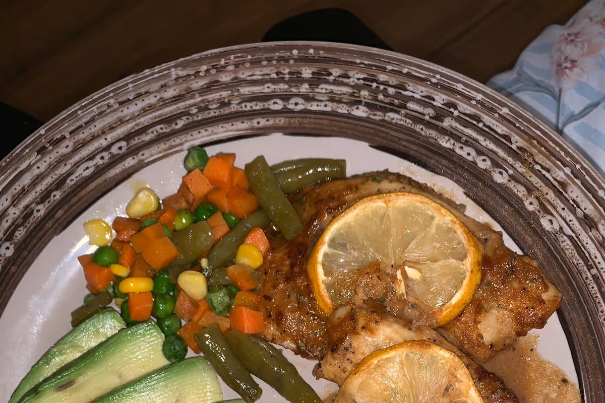 A close-up view of a plate with cooked chicken topped with lemon slices, served with mixed vegetables including green beans, peas, carrots, and corn, and slices of zucchini on the side.