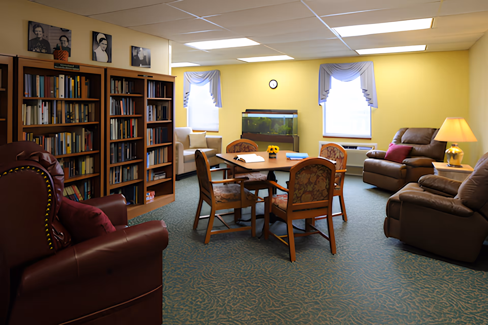 A cozy common room with yellow walls featuring a round wooden table surrounded by four cushioned chairs in the center. There are two large bookshelves filled with books on the left side, a fish tank against the far wall between two windows with blue valances, and several comfortable armchairs and recliners arranged around the room. A table lamp is lit on a side table next to one of the recliners.