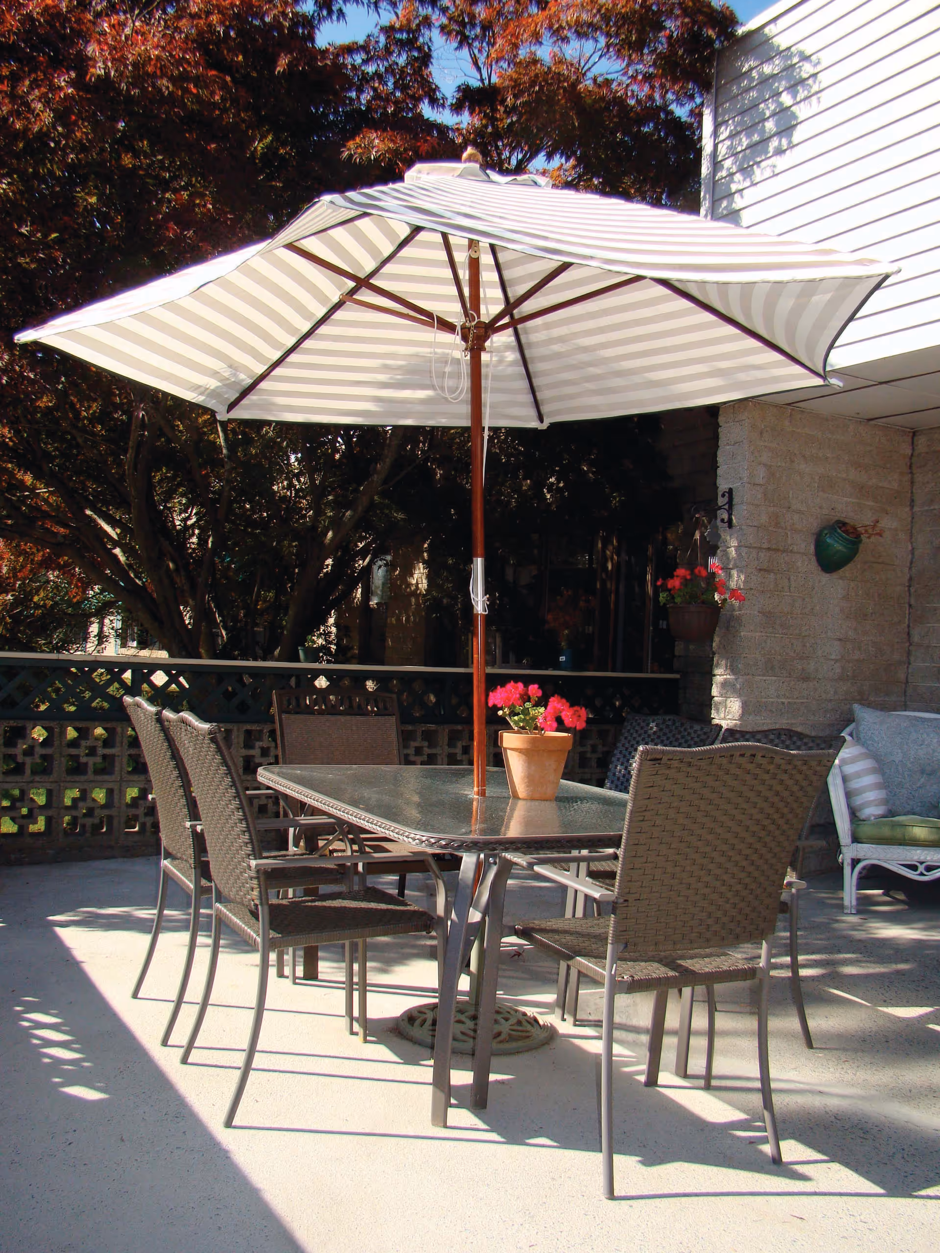 Outdoor patio area with a glass-top table surrounded by six wicker chairs. A large striped umbrella provides shade over the table, which has a small potted plant with red flowers in the center. In the background, there are trees with autumn-colored leaves and part of a building with a stone wall and a cushioned chair.