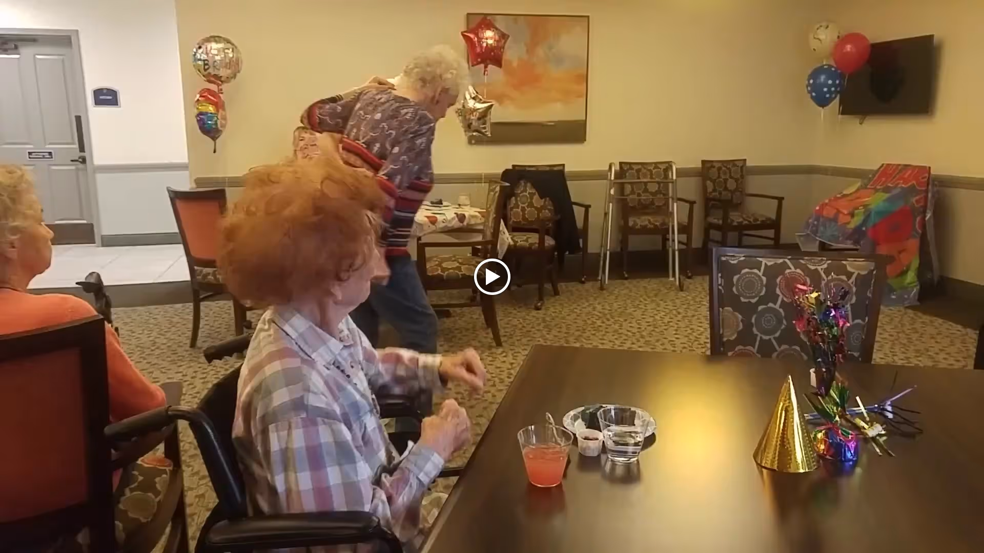 A group of elderly women in a decorated common room with balloons and party decorations. One woman with curly red hair is seated at a table with a party hat, a drink, and small bowls. Other women are seated or standing in the background near chairs and a walker.