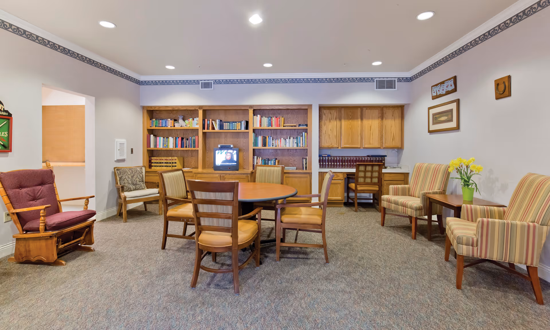 A cozy common area in a senior living facility with a round wooden table surrounded by four chairs in the center. To the left, there is a wooden rocking chair with a maroon cushion and another chair with a patterned cushion. The back wall features built-in wooden bookshelves filled with books and a small TV. To the right, there is a small desk with a chair and wooden cabinets above it. Two striped armchairs with a small wooden table between them, holding a green vase with yellow flowers, are placed against the right wall. The room has beige walls with a decorative border near the ceiling and carpeted flooring.