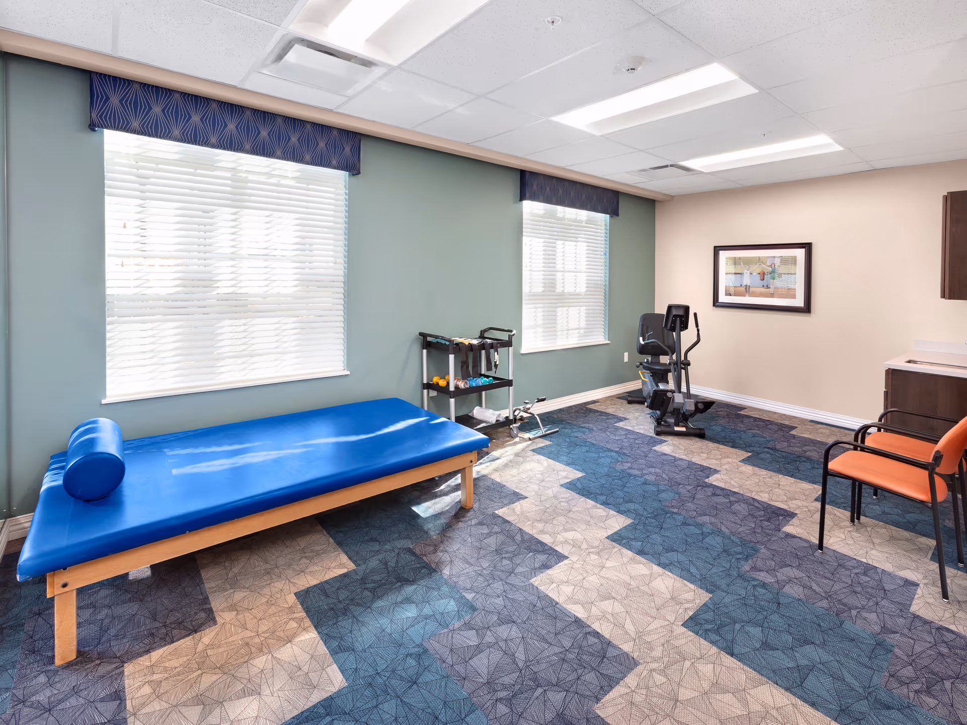 A bright therapy/exercise room with a blue treatment table, exercise bike, small weights rack, and chairs.
