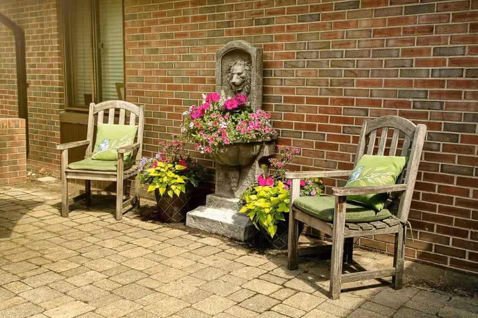 Outdoor patio area with two wooden chairs featuring green cushions and pillows, a stone lion head water fountain adorned with pink flowers, and two potted plants with yellow and pink flowers against a brick wall.