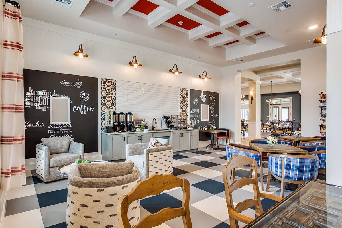 A bright and inviting common area with a coffee station featuring multiple coffee dispensers and a variety of coffee supplies. The room has a checkered black and white floor, comfortable armchairs, and wooden tables with blue plaid cushioned chairs. The ceiling has a white grid design with red panels, and the walls have decorative blackboards with coffee-themed writings and illustrations. The space opens into a dining area with more tables and chairs.