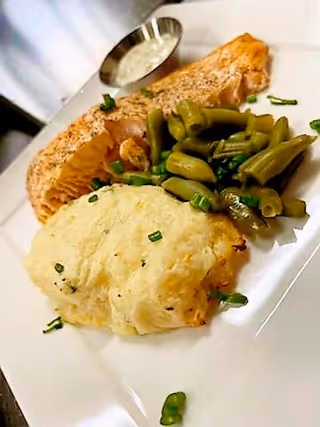 A plated meal consisting of a piece of cooked salmon, a serving of green beans, and a portion of mashed potatoes garnished with chopped chives, with a small container of white sauce on the side.