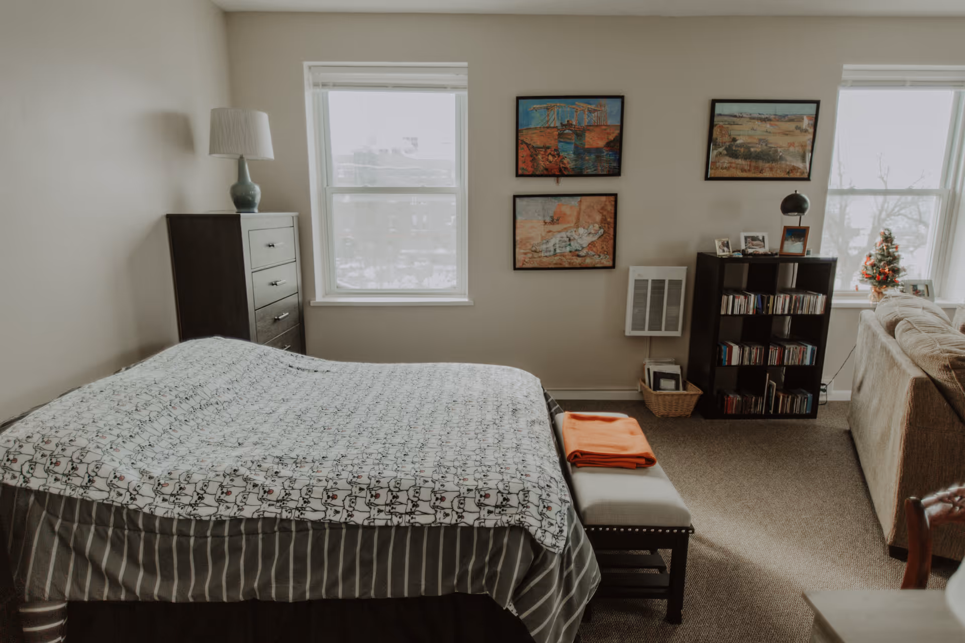 A cozy bedroom with a bed covered in a patterned blanket and striped bed skirt. There is a dark wooden chest of drawers with a lamp on top to the left, and two windows letting in natural light. On the right wall, there are two framed paintings above a small heater. A black bookshelf filled with CDs and framed photos sits next to a beige couch, with a small decorated Christmas tree on the windowsill.