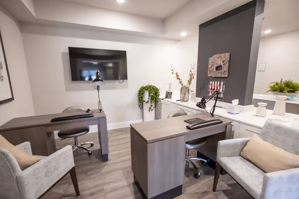 A modern office or consultation room with two desks and chairs, a wall-mounted flat screen TV, decorative plants, and neutral-toned armchairs with beige cushions. The room has wood flooring and white walls with a dark accent panel displaying small decorative items.