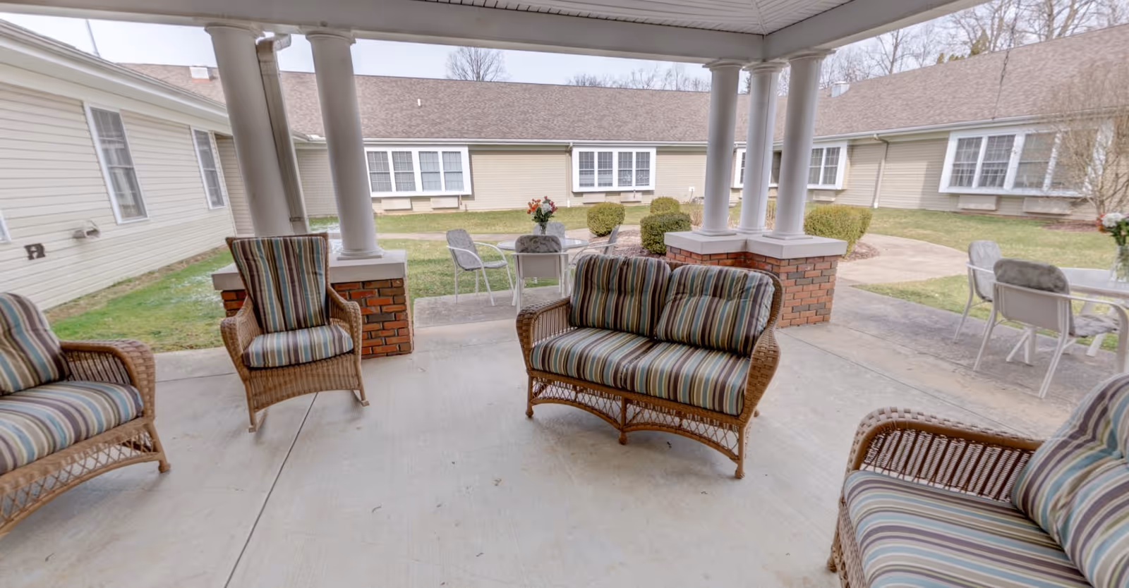 Covered outdoor patio area with wicker furniture including a loveseat and chairs with striped cushions. The patio overlooks a grassy courtyard surrounded by single-story beige buildings with multiple windows. There are also small tables and chairs with flower arrangements on them in the courtyard.