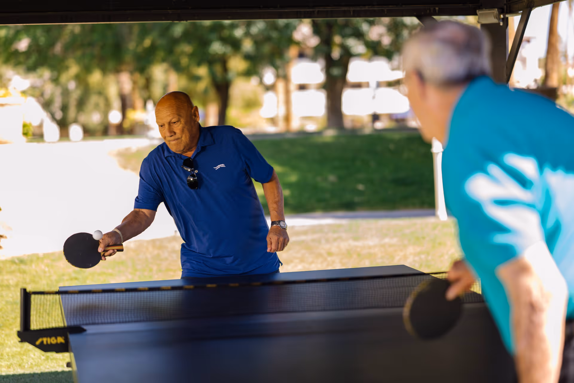 Two elderly men playing table tennis outdoors under a shaded area with green grass and trees in the background. One man is wearing a blue shirt and is about to hit the ball, while the other man, wearing a teal shirt, is preparing to return the shot.