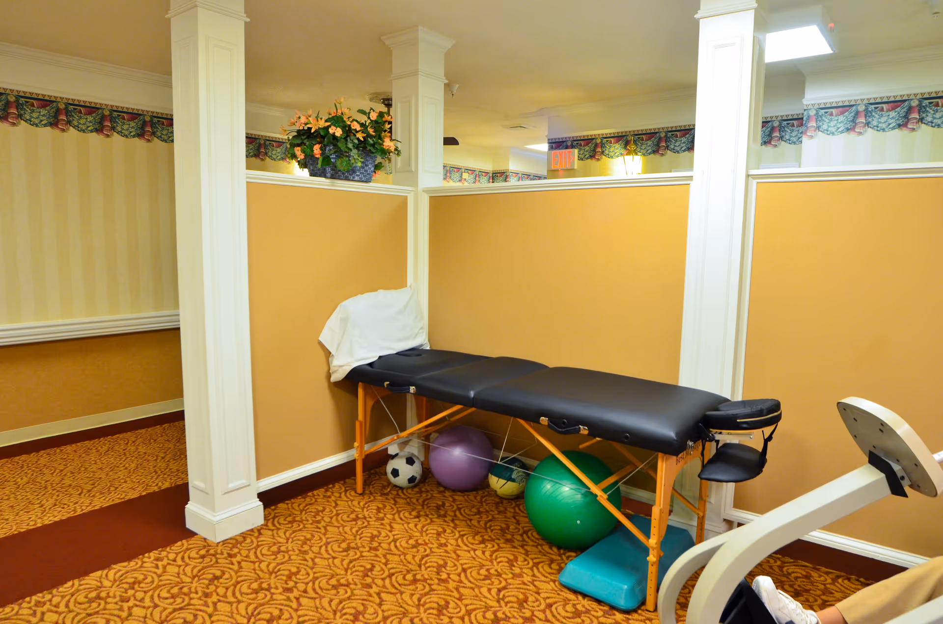 A therapy or massage table with a white towel draped over one end, positioned against a beige partition wall in a room with patterned carpet and wallpaper. Underneath the table are various exercise balls and a soccer ball. Part of an exercise machine is visible on the right side of the image.