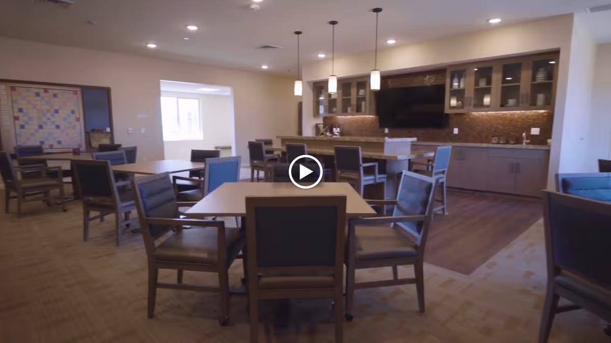 Open communal dining and activity room with multiple tables and chairs, a kitchen counter with pendant lights, and a wall-mounted TV.