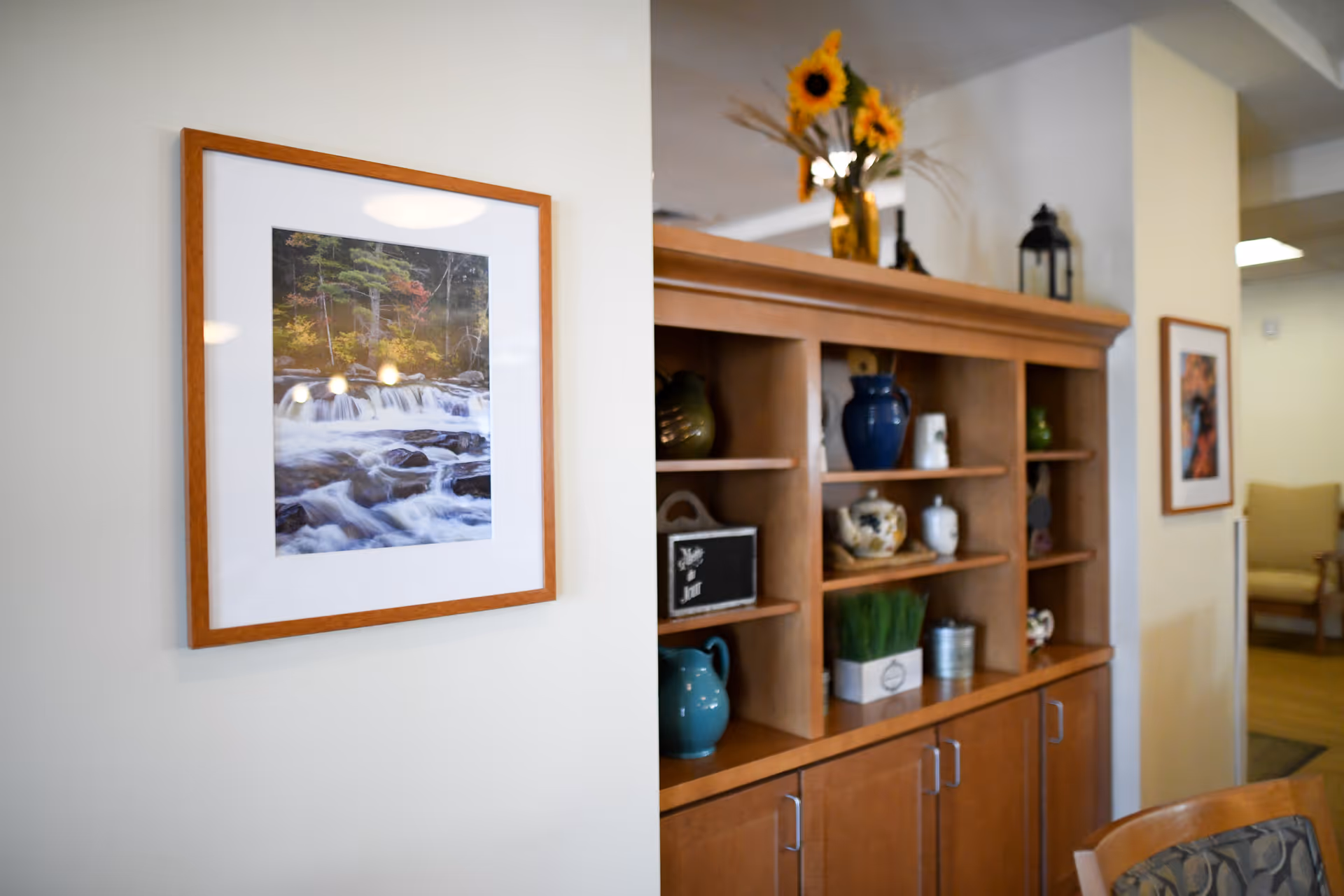 Interior view of a senior living facility showing a wooden shelving unit with various decorative items including vases, a small chalkboard, and a plant. On the wall to the left, there is a framed photograph of a flowing river with trees in the background. In the background, there is a glimpse of a seating area with a chair and another framed picture on the wall.